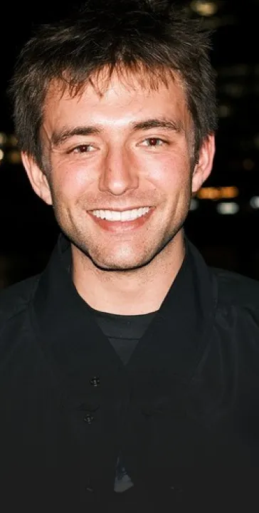 Young man with dark tousled hair smiling against a dark background.